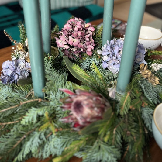 Tannengrüner Adventskranz mit vier grünen Kerzen, dekoriert mit getrockneten Hortensienblüten und weiteren Trockenblumen; im Hintergrund steht eine Teetasse.